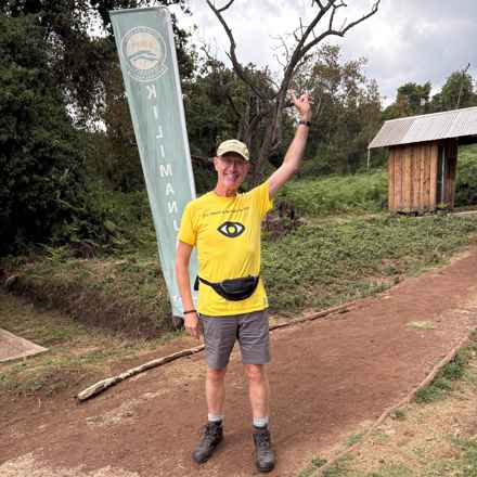 Kevin dressed in his yellow Macular Society t-shirt, smiling with one hand in the air, as he prepares to take on Mount Kilimanjaro
