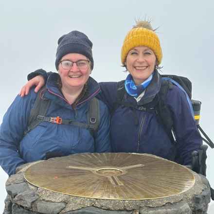 Shenay, left, and Cath smiling as they pose for a quick mountain summit photo. Cath is wearing a grey hat, glasses and navy blue jacket with black backpack. Cath is wearing a mustard yellow hat, dark purple coat with scarf and black rucksack.